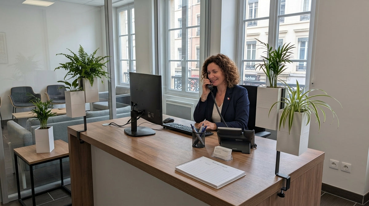 Une femme de bureau portant un blazer bleu parle au téléphone à son poste de travail. Son bureau est équipé de deux supports d'îlot végétal noirs à pince de la marque Éclance, chacun tenant deux cache-pots 'Le cube' blancs de 6cm, plantés de fougère, palmier et plantes araignées. Le fond montre un bureau ouvert avec des cloisons vitrées, des canapés et des fenêtres donnant sur un bâtiment parisien.