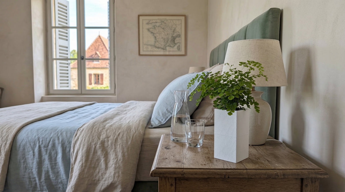 Cache-pot Éclance Le Cube blanc (pot 6cm) avec une fougère Adiantum, posé sur une table de chevet en bois brut dans une chambre d'hôtes lumineuse.
