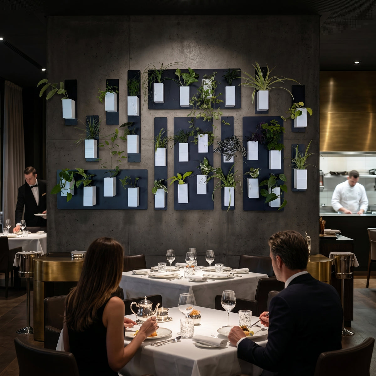 Une vue élégante de la salle d'un restaurant gastronomique. Un couple en tenue de soirée dîne au premier plan devant un mur texturé effet béton ciré. Le point focal est une vaste installation murale Éclance décoration intérieur végétal, composée de nombreux cache-pots Le cube blancs, parfaitement adaptés pour un pot de 6cm, fixés sur des supports noirs et élégamment rétroéclairés. L'arrière-plan révèle un serveur et un chef dans une cuisine ouverte.