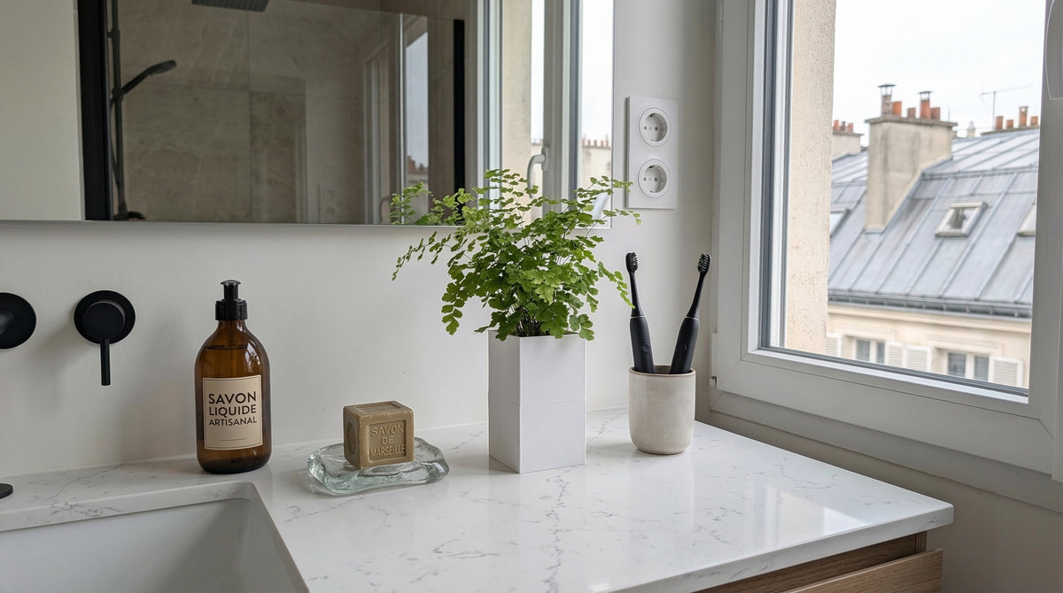 Cache-pot Éclance Le Cube blanc pour pot de 6cm avec une fougère, disposé sur un plan vasque en marbre blanc devant une fenêtre avec vue sur les toits de Paris.