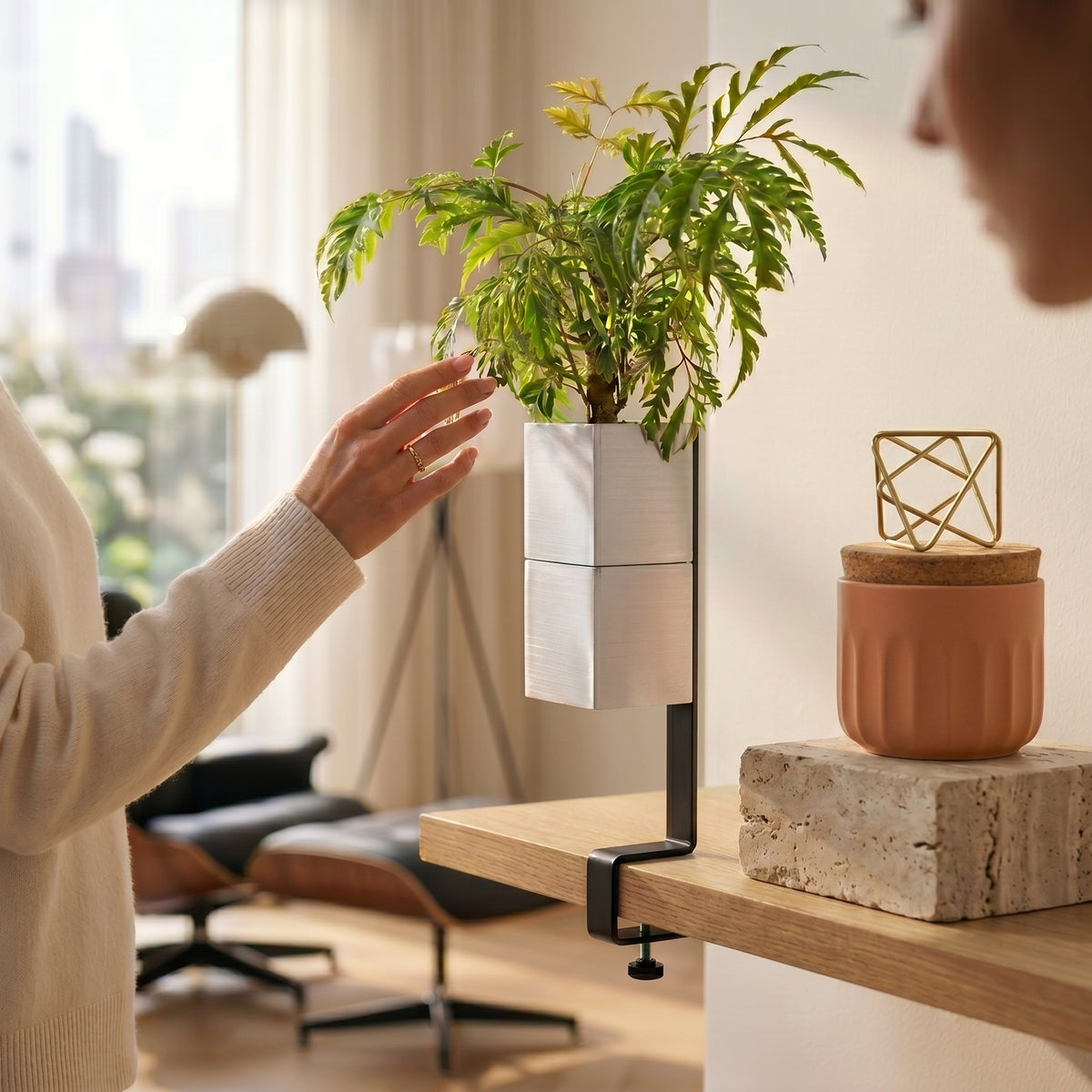 Cache-pot Le cube couleur blanc de la marque Éclance décoration intérieur végétal, adapté pour un pot de 6cm. Il est suspendu à une étagère en bois grâce à un élégant support étau noir ilot végétal,  mettant en valeur une plante verte d'intérieur touchée délicatement par une femme dans un salon moderne.