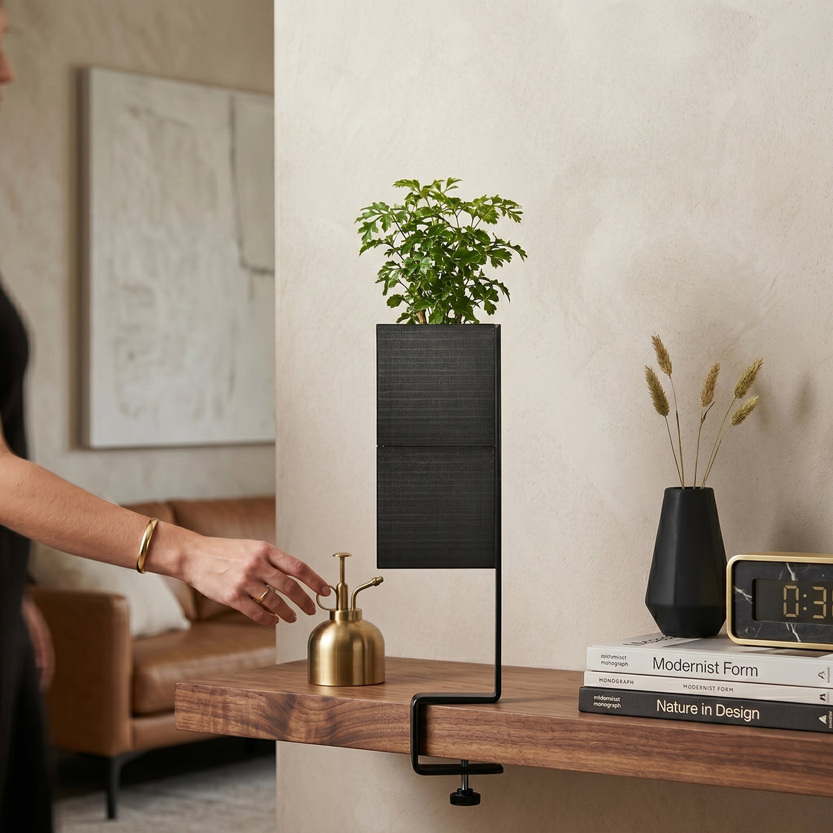 L'îlot végétal avec son cache-pot Le cube de couleur noire de la marque Éclance décoration intérieur végétal, adapté pour un pot de 6cm. Accueillant une petite plante verte, il est fixé au bord d'une étagère en bois foncé grâce à son support étau noir. Dans ce décor de salon au design moderne, une main féminine s'approche d'un petit vaporisateur doré.