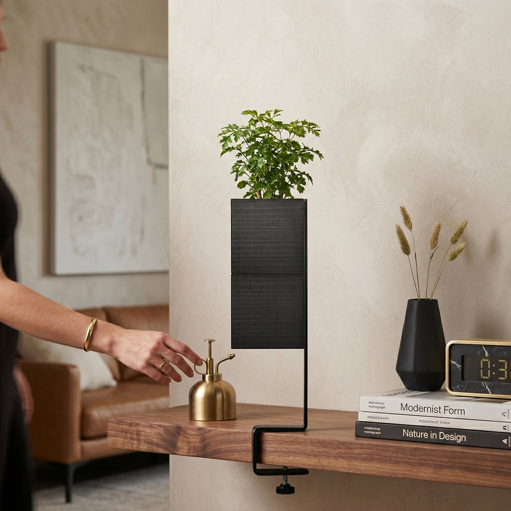 L'îlot végétal avec son cache-pot Le cube de couleur noire de la marque Éclance décoration intérieur végétal, adapté pour un pot de 6cm. Accueillant une petite plante verte, il est fixé au bord d'une étagère en bois foncé grâce à son support étau noir. Dans ce décor de salon au design moderne, une main féminine s'approche d'un petit vaporisateur doré.