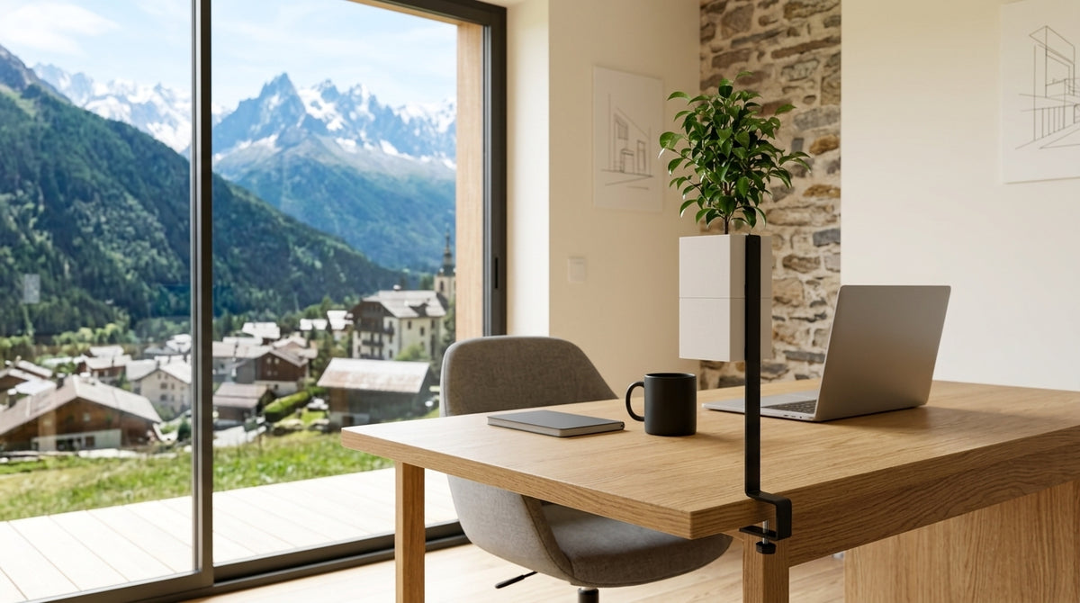 Support îlot végétal Éclance noir avec son cache-pot Le Cube blanc (pour pot de 6cm) fixé à un bureau en bois. L'espace de travail lumineux fait face à une grande baie vitrée révélant un village alpin et des montagnes enneigées.