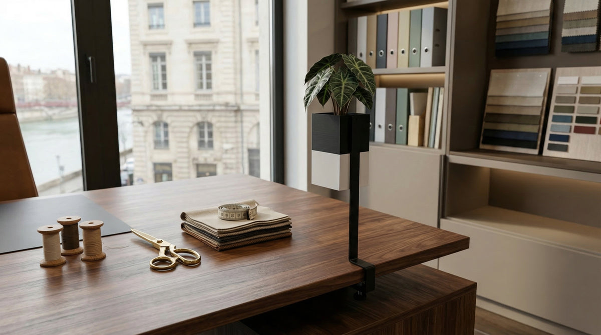Cache-pot bicolore noir et blanc de la marque Éclance, modèle "Le cube" (adapté pour un pot de 6cm), fixé au bord d'un élégant bureau en bois grâce à son support pince "l'îlot végétal". Il met en valeur une petite plante verte (Ficus Lyrata). Sur le bureau sont disposés de manière ordonnée un sous-main, un plateau avec des échantillons de tissus carrés, un pot à stylos et une lampe design en laiton.