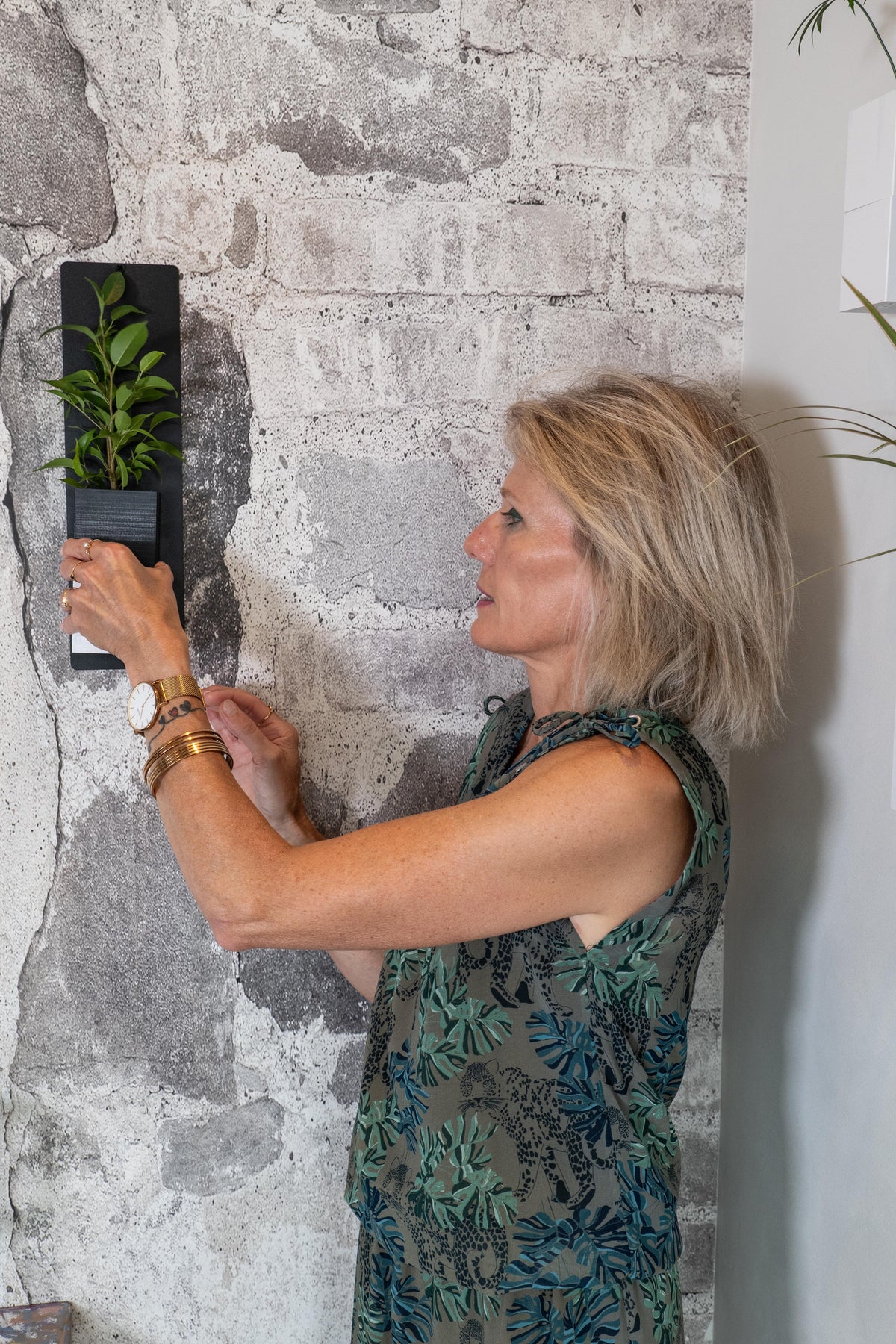 Mini mur végétal Éclance M1 bicolore (cube haut noir, cube bas blanc) : une femme installe un mini support mural avec une plante verte sur un mur en pierre.