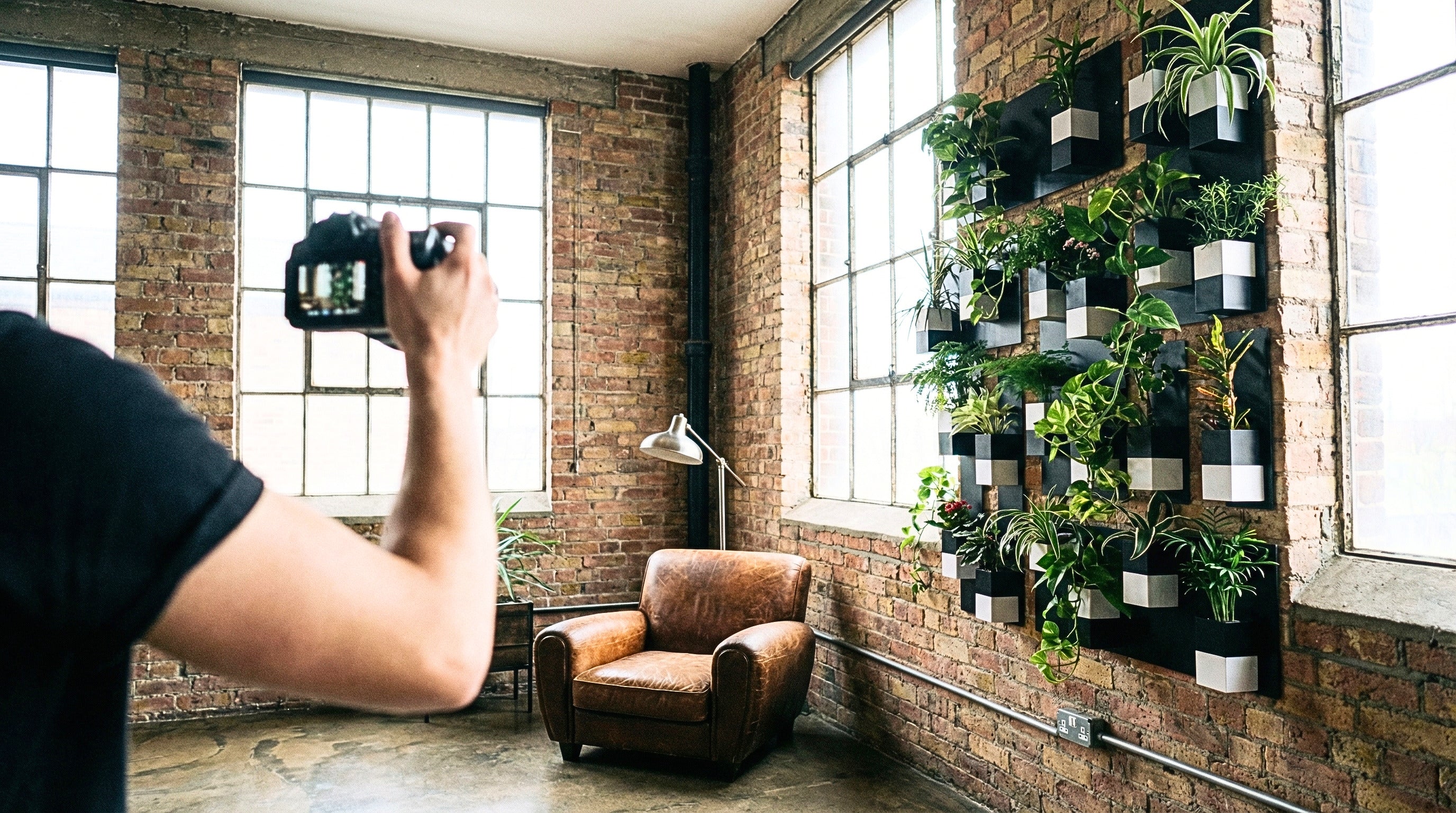Un photographe prend en photo un grand mur végétal intérieur modulaire d'Éclance. Ce mur est composé de nombreux cache-pots "Le Cube" bicolores (noir et blanc), chacun étant un cache-pot adapté pour un pot de 6cm, rempli de plantes variées (Pothos, fougères, palmiers nains). L'installation est fixée sur un mur de briques apparentes dans un salon de style loft industriel, à côté d'un fauteuil club en cuir marron et de grandes fenêtres. Le cache-pot Le Cube 6cm est au 